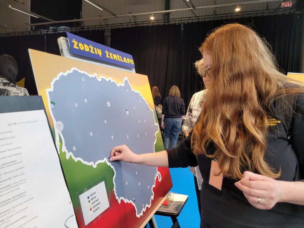 Moteris stovi prie stendo su Lietuvos žemėlapiu ir smeigia smeigtuką į pasirinktą vietą. Viršuje matyti užrašas „Žodžių žemėlapis“, aplink – parodos erdvė su kitais žmonėmis.