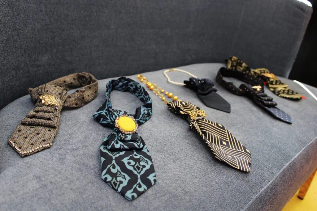 Ant pilkos kėdės išdėstyti keli dekoratyviniai kaklo papuošalai, primenantys kaklaraiščius – skirtingų spalvų ir raštų, su aukso spalvos detalėmis ir pakabukais.