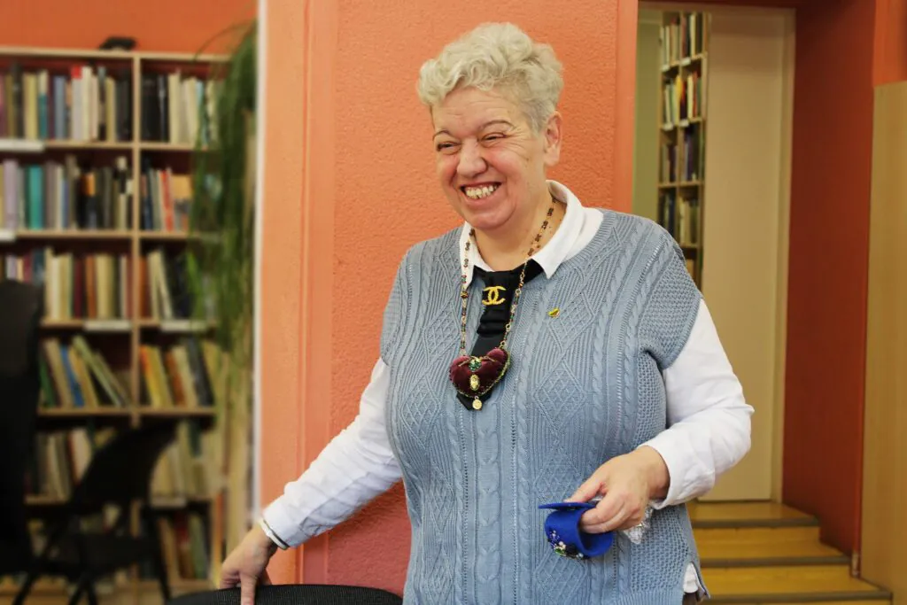 Bibliotekos erdvėje stovi besijuokianti moteris su pilka megzta liemene ir baltais marškiniais, seginti didelį rankų darbo kaklo papuošalą su širdelės formos pakabuku.