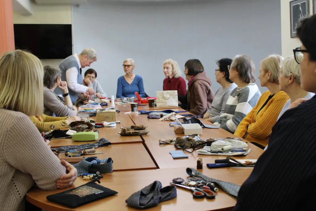 Kūrybinių dirbtuvių metu žmonių grupė sėdi prie stalo, klausosi vedėjos ir kuria rankų darbo kaklo papuošalus iš įvairių medžiagų.
