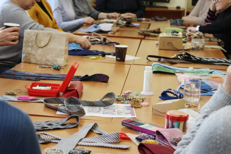 Žmonės sėdi prie didelio stalo ir užsiima rankdarbiais – ant stalo išdėlioti kaklaraiščiai, siūlai, žirklės, sagos, papuošalų detalės ir kitos kūrybinės priemonės.