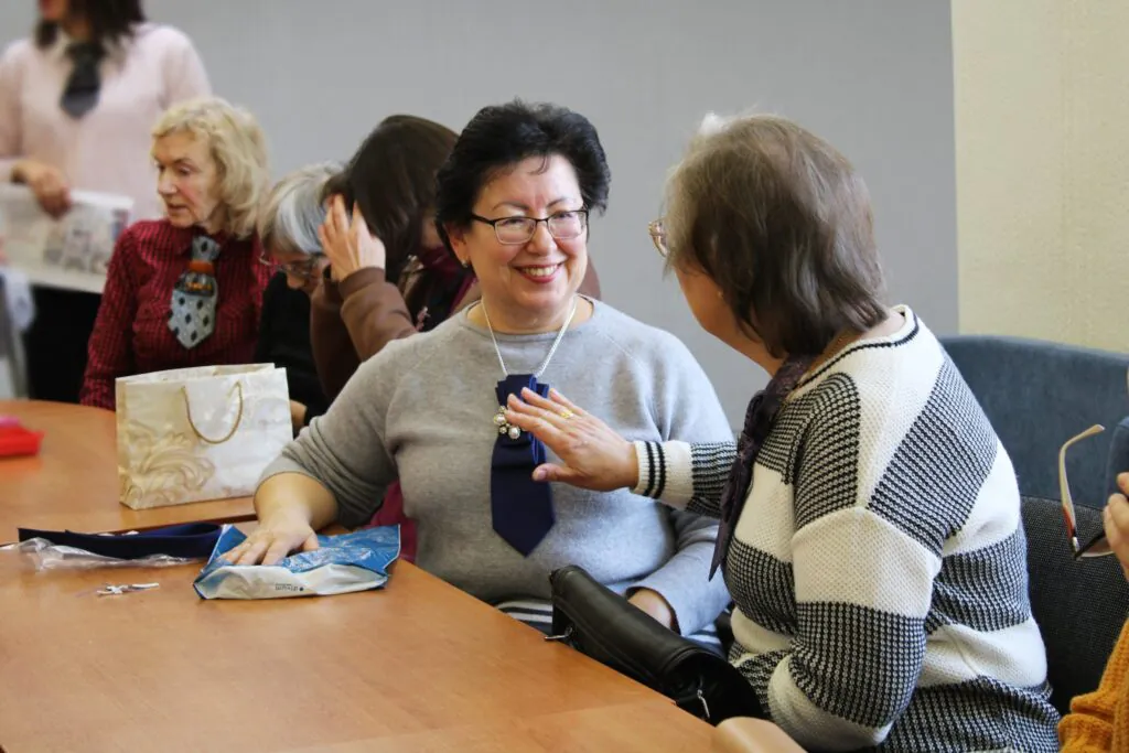 Dvi moterys sėdi prie stalo ir bendrauja kūrybinių dirbtuvių metu – viena jų ant megztinio prisisegusi iš kaklaraiščio pagamintą papuošalą, o aplink matyti kiti dalyviai ir rankdarbių priemonės.