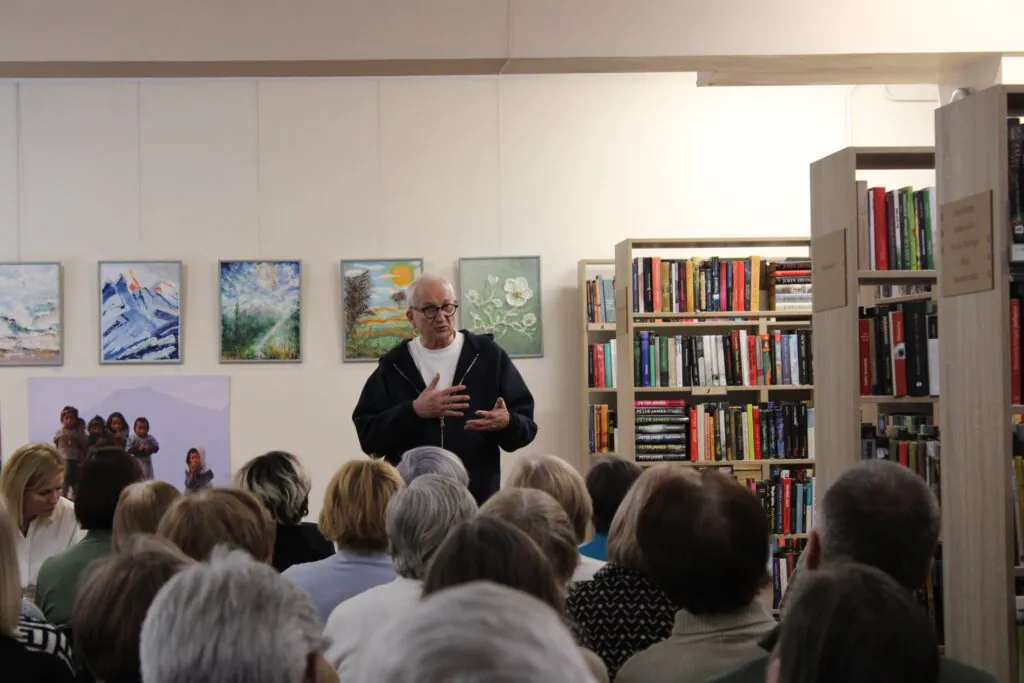 Nuotraukoje matomas vyras, kalbantis prieš auditoriją, stovintis bibliotekoje. Už jo yra nuotraukos ir meno darbai, o jis pats gestikuliuoja rankomis, aiškindamas temą. Pirmame plane matoma klausytojų grupė. Jų žvilgsniai nukreipti į kalbėtoją.