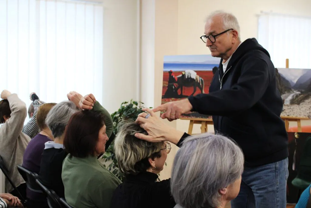 Nuotraukoje matomas vyras, kalbantis su auditorija ir rodantis į vieną iš klausytojų, kuriam jis taiko praktinę demonstraciją. Moteris, sėdinti pirmoje eilėje, laiko savo galvą, o kitos klausytojos taip pat seka instrukcijas. Už jų matoma nuotrauka su žmonėmis, vedančiais arklius per raudoną žemę.