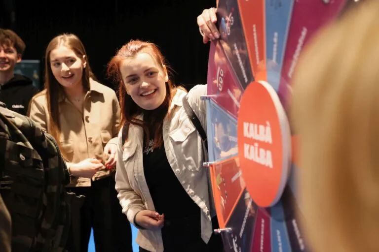 Trys besišypsantys jauni žmonės stovi prie žaidimo rato, viena mergina jį suka